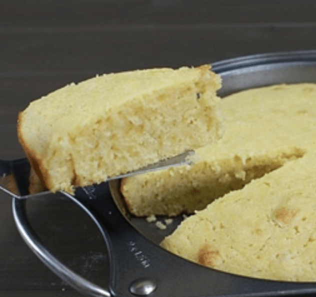 A slice of cornbread, made from a classic cornbread recipe, is being lifted from a round metal pan with a spatula.