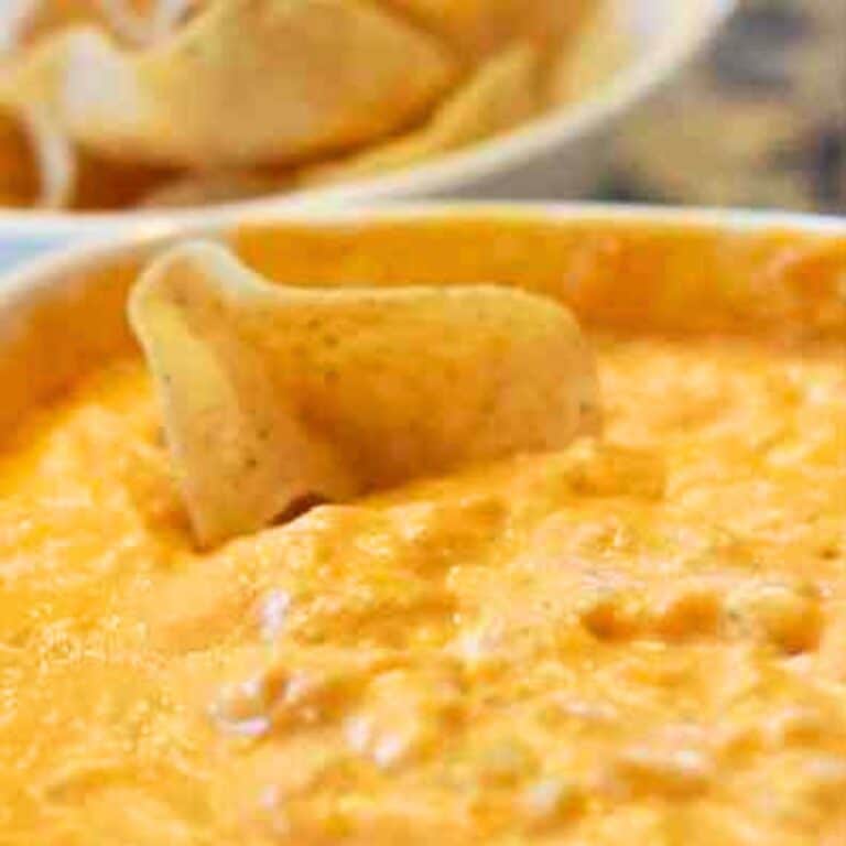 A tortilla chip is dipped into a bowl of creamy, orange-colored cheese dip—an easy buffalo chicken dip—while another bowl of chips sits invitingly in the background.
