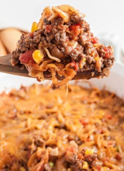 A wooden spoon holds a serving of cheesy hamburger casserole with ground beef, noodles, corn, tomatoes, and melted cheese above a white baking dish.