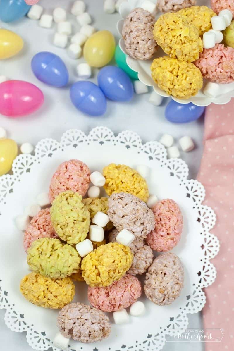 crispy treat eggs, yellow, pine and light purple on white tray surrounded by Easter eggs and mini-marshmallows
