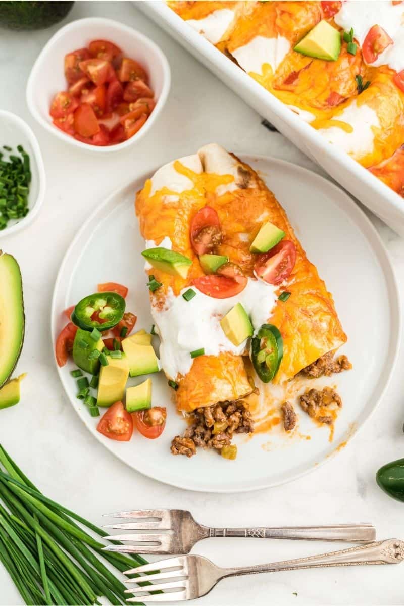 two enchiladas on plate with sour cream, tomatoes, and peppers on top, two forks on the side