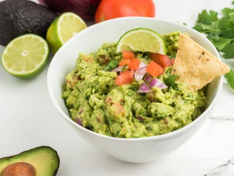 white bowl with guacamole topped with lime slice, diced tomatoes and onion with two chips
