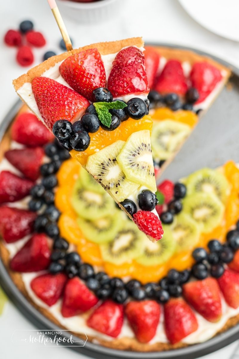 fruit pizza with slice cut out: strawberries, blueberries, mandarin oranges, kiwi, blueberries, raspberries in the middle. 
