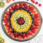 fruit pizza with slice cut out: strawberries, blueberries, mandarin oranges, kiwi, blueberries, raspberries in the middle.