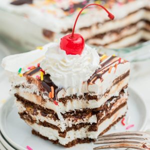 slice of ice cream sandwich cake. You can see layers of chocolate wafers and vanilla ice cream, topped with sprinkles, whipped cream and a cherry with a stem
