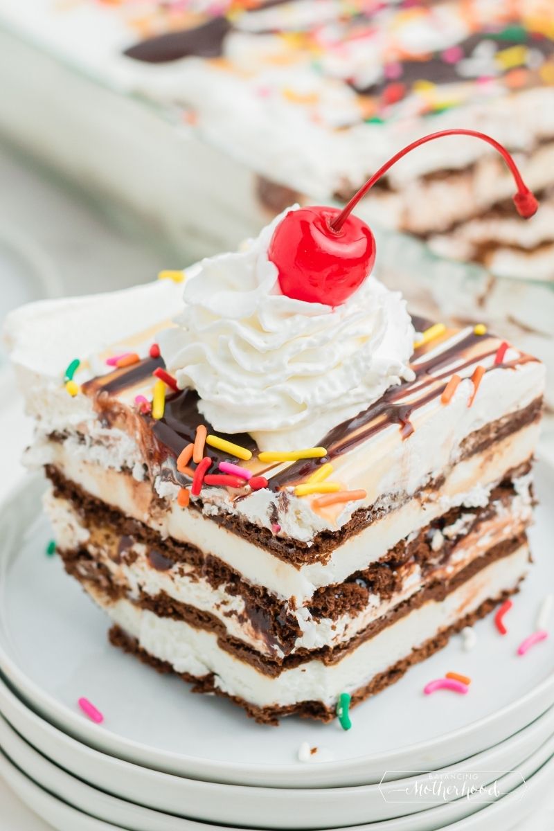 slice of ice cream sandwich cake. You can see layers of chocolate wafers and vanilla ice cream, topped with sprinkles, whipped cream and a cherry with a stem