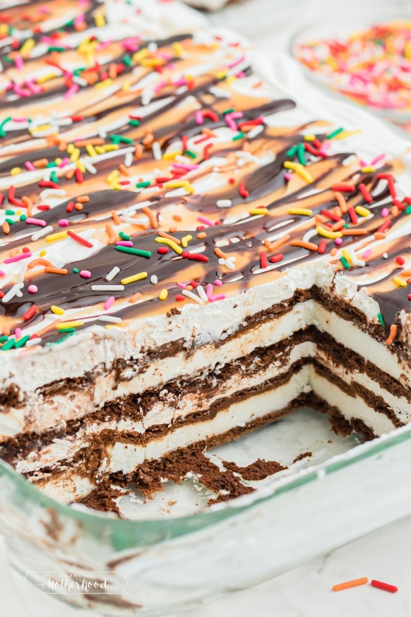 slice of ice cream sandwich cake. You can see layers of chocolate wafers and vanilla ice cream, topped with sprinkles, whipped cream and a cherry with a stem
