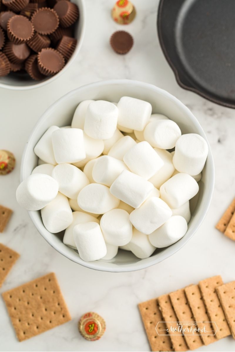 white bowl of large marshmallows, white bowl with unwrapped mini reese's candies, black skillet