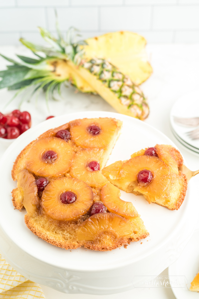 round cake on white plate. Golden pineapple rings with a cherry in the middle of each one, and a slice taken out with whole pineapple in background

