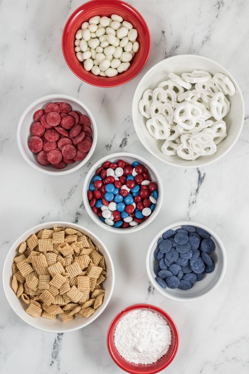 ingredients in bowls on white counter: red, white, blue candy melts and M&Ms, chex mix cereal, powdered sugar, white coated pretzels