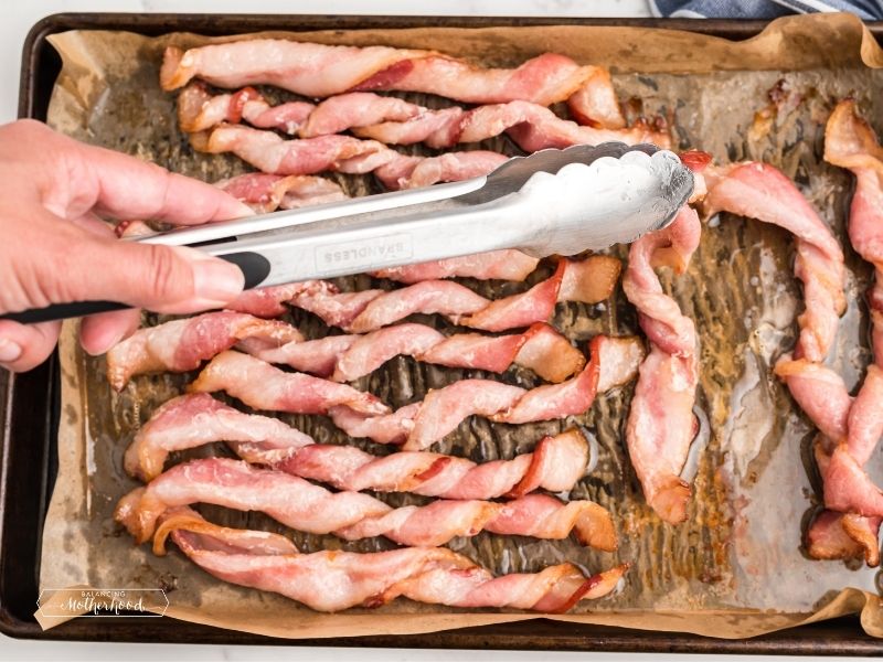 bacon twisted on baking sheet with parchment paper. Tongs flipping bacon