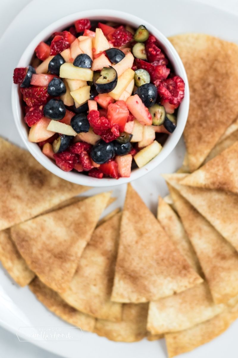 white bowl with mixed berry fruit salsa (melon, raspberries, grapes, apple) with triangle-shaped cinnamon chips on the side