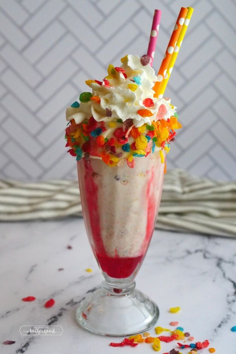 milkshake glass with strawberry syrup on inside, whipped cream and fruity pebbles cereal on top