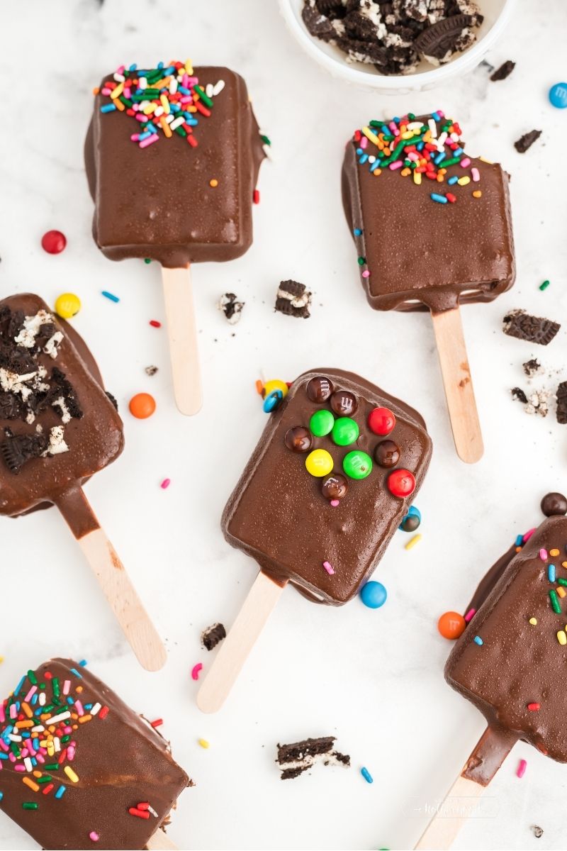 ice cream sandwiches covered in chocolate with popsicle stick and sprinkles laying on white table