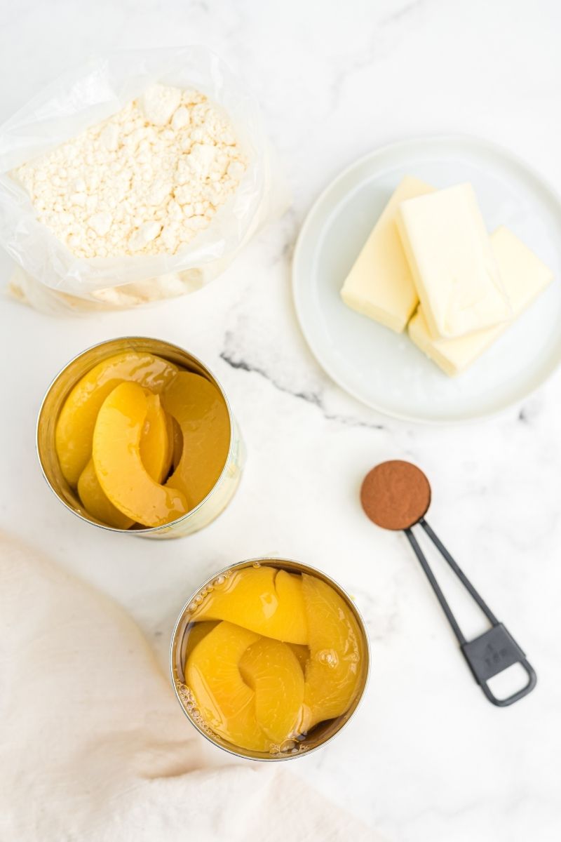 ingredients on white counter: bag of white cake mix, 3 small sticks of butter, two open cans of peaches, measuring spoon with cinnamon