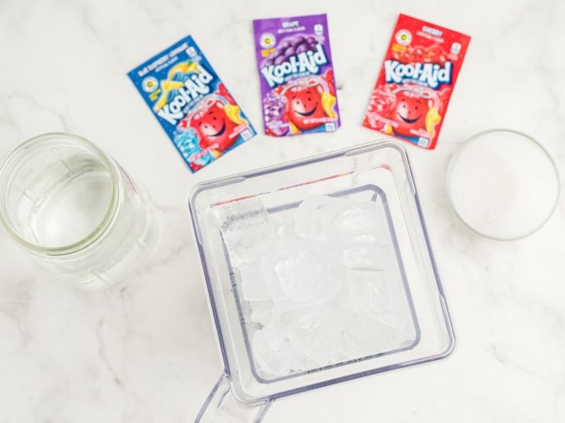 white counter with ingredients: three Kool-Aid packets, blue, purple and red, bowl of sugar, blender with ice, mason jar with water