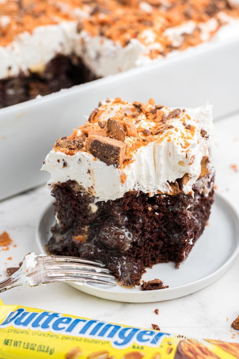 square piece of chocolate cake with whipped cream and butterfinger candy pieces on top with fork on the side