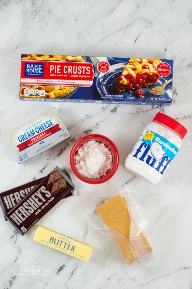 ingredients on white marble counter: pie crust, cream cheese, powdered sugar in red bowl, marshmallow fluff jar, 2 Hershey's candy bars, pack of graham crackers, one stick of butter