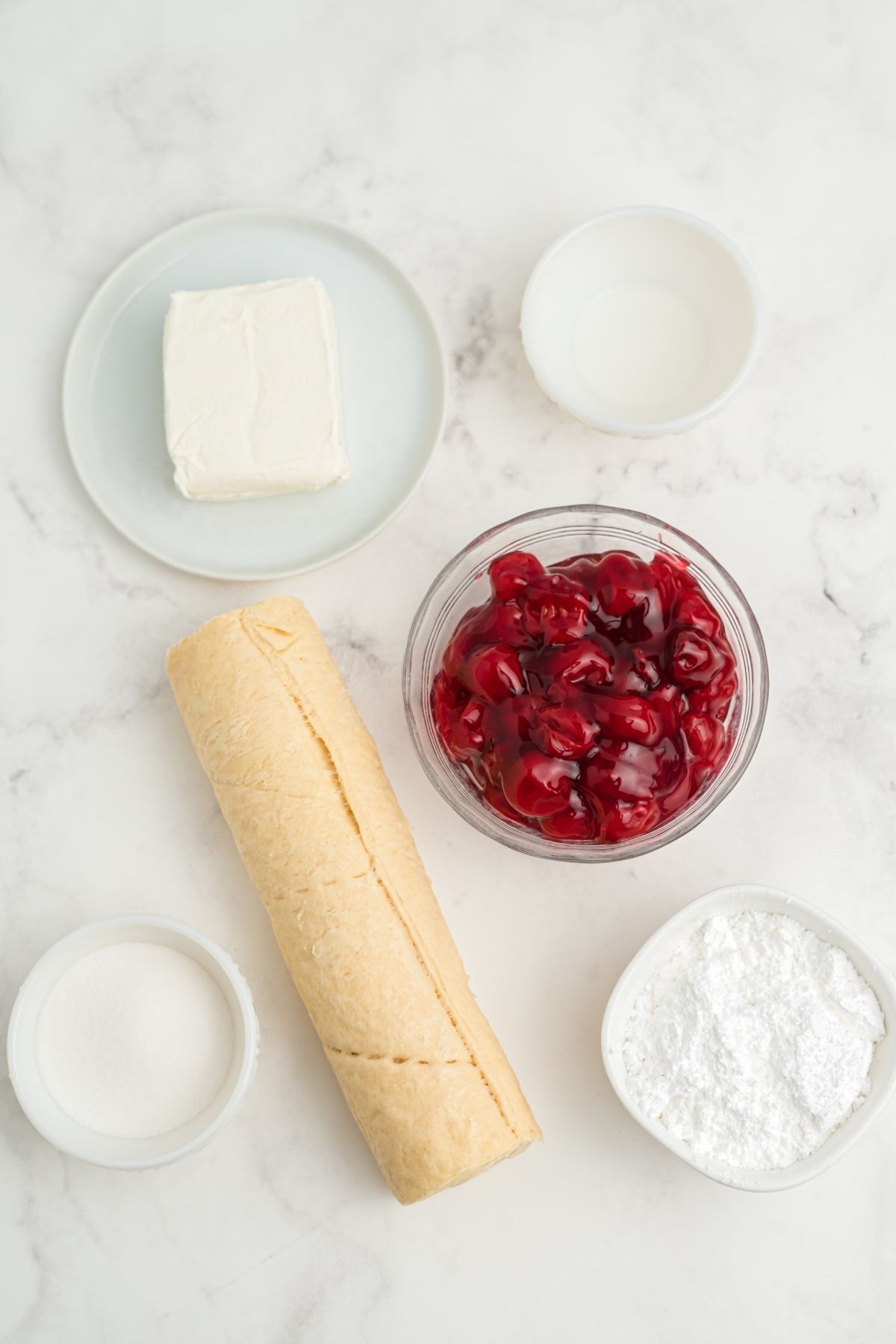 ingredients on white marble counter: block of cream cheese, milk, cherry pie filling, powdered sugar, rolled crescent roll dough, sugar