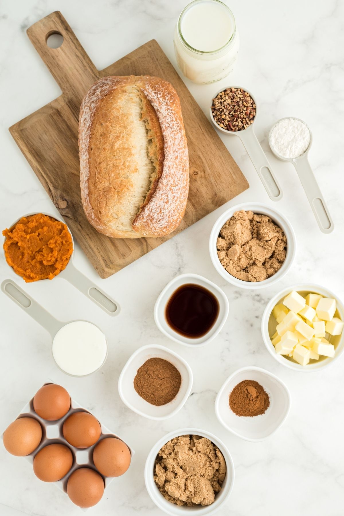 ingredients on white counter: loaf of crusty French bread, pumpkin puree, brown eggs, diced butter, vanilla, pumpkin pie spice, heavy cream, and milk