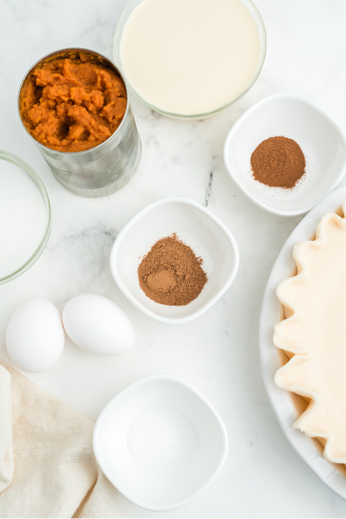 Classic Pumpkin Pie ingredients on a white counter. The ingredients are the following: sugar, cinnamon, salt, pumpkin pie spice blend, large eggs, pumpkin, evaporated milk, and deep dish pie shell. 
