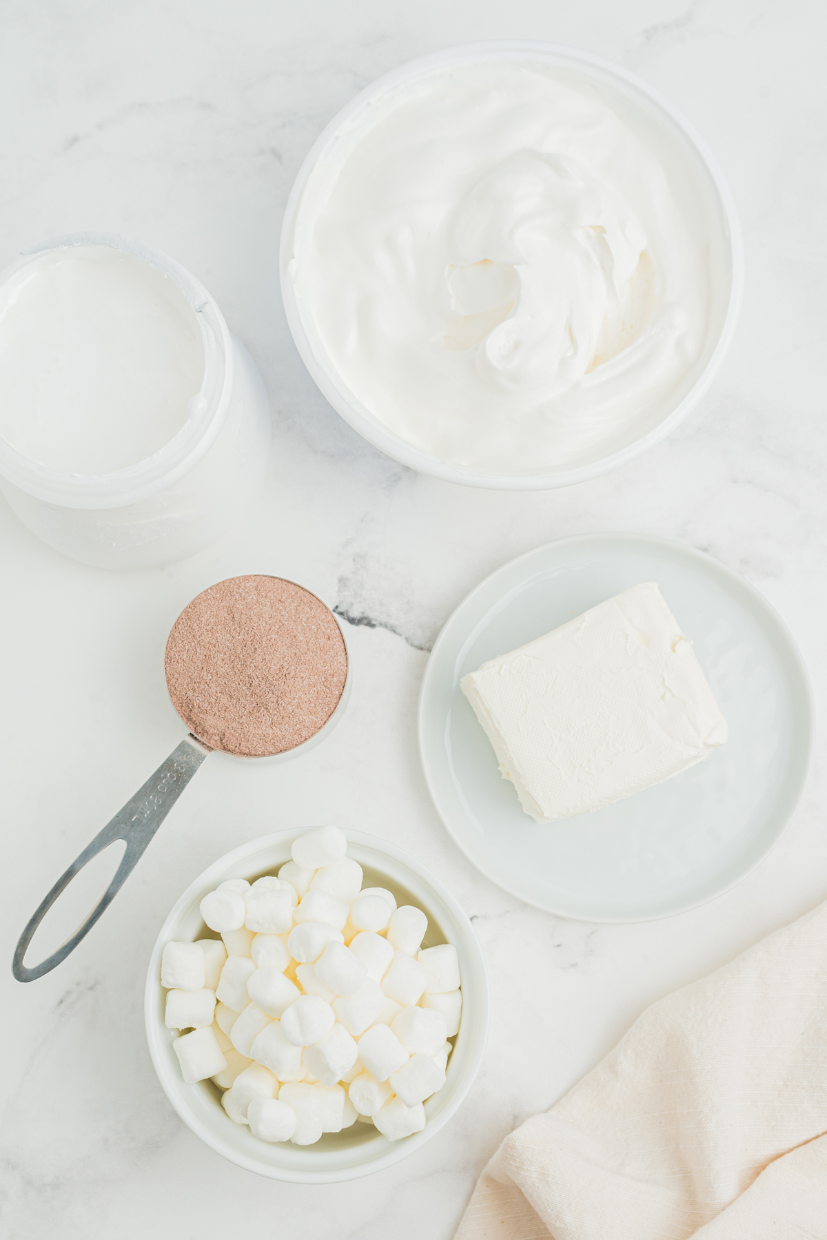 Hot chocolate dip Ingredients on a white counter. These are marshmallow fluff, cream cheese hot chocolate mix, whipped topping, and thawed mini marshmallows for garnish.