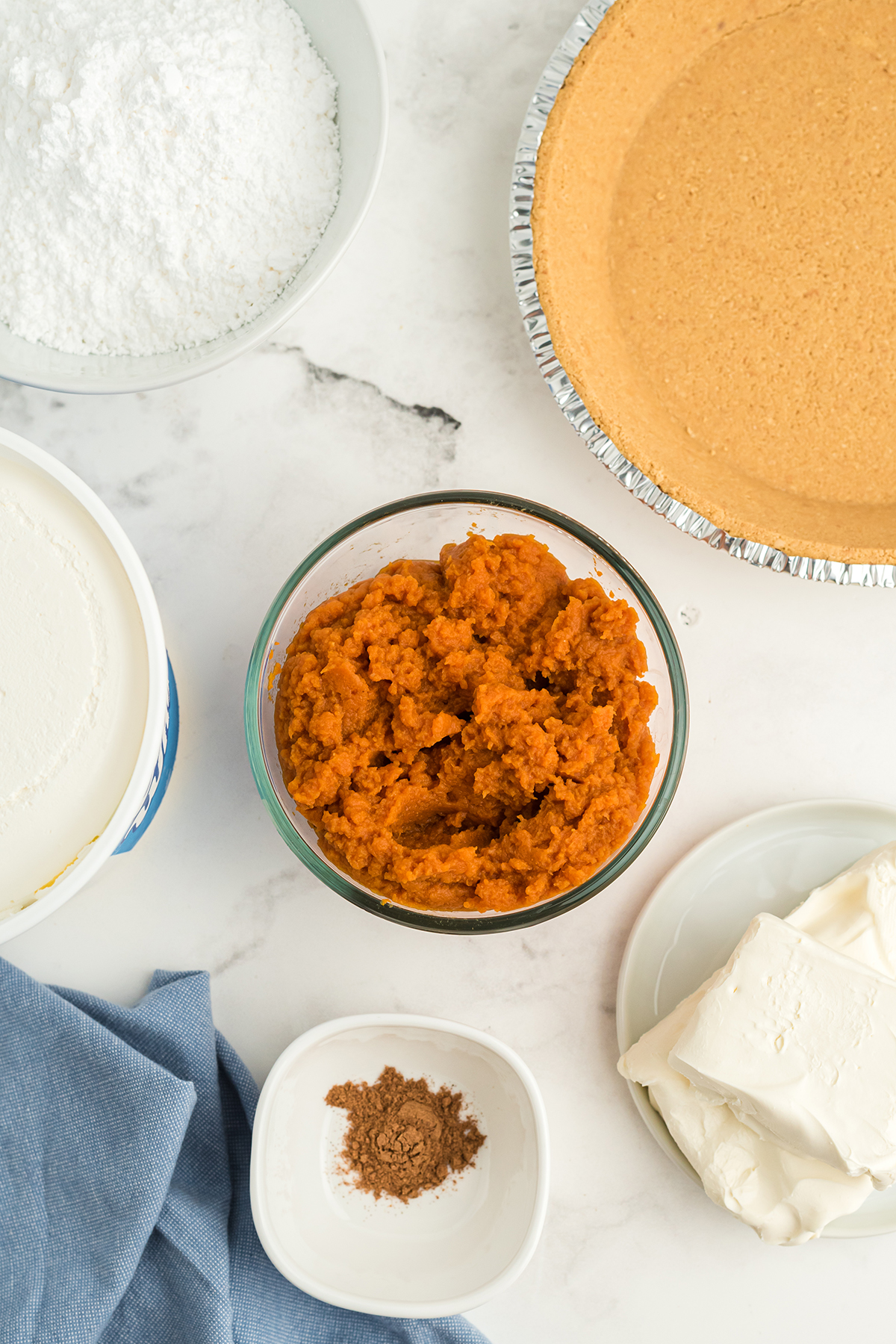 Cool Whip Pumpkin Pie ingredients on a white counter with a blue linen. The ingredients are pumpkin puree, milk, graham cracker crust, whipped cream, and vanilla pudding.