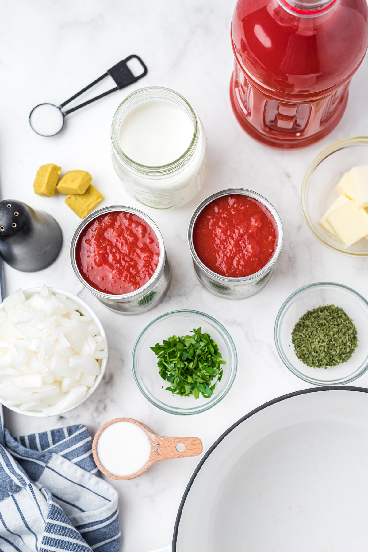 Ingredients of tomato soup on a white counter. These are the following: onion, butter, crushed tomatoes, tomato juice, sugar, chicken bouillon cubes, heavy cream, parsley, dried basil, salt, and pepper.
