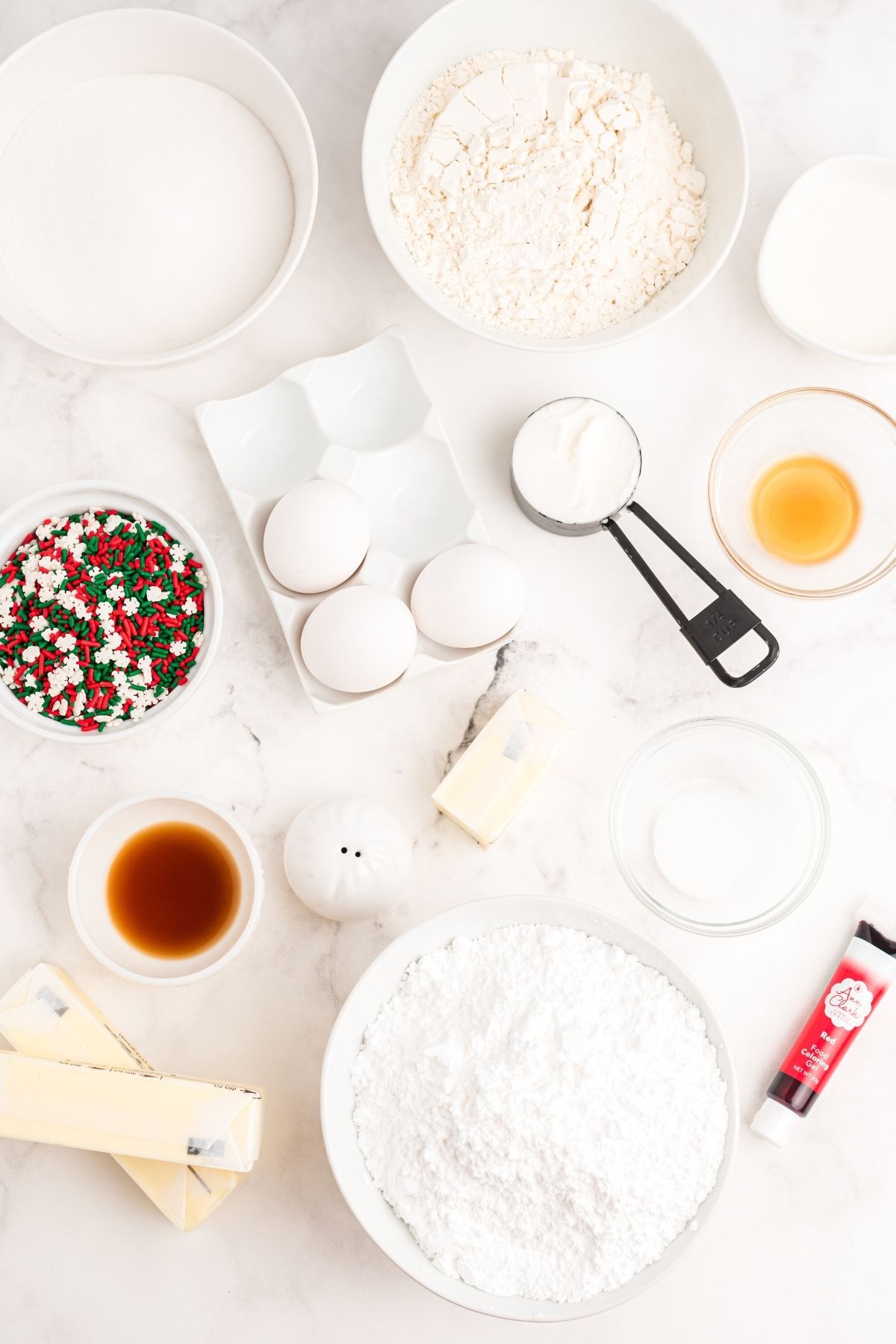 ingredients on white counter: butter, sugar, 3 white eggs, vanilla, Christmas sprinkles, powdered sugar, food coloring, milk