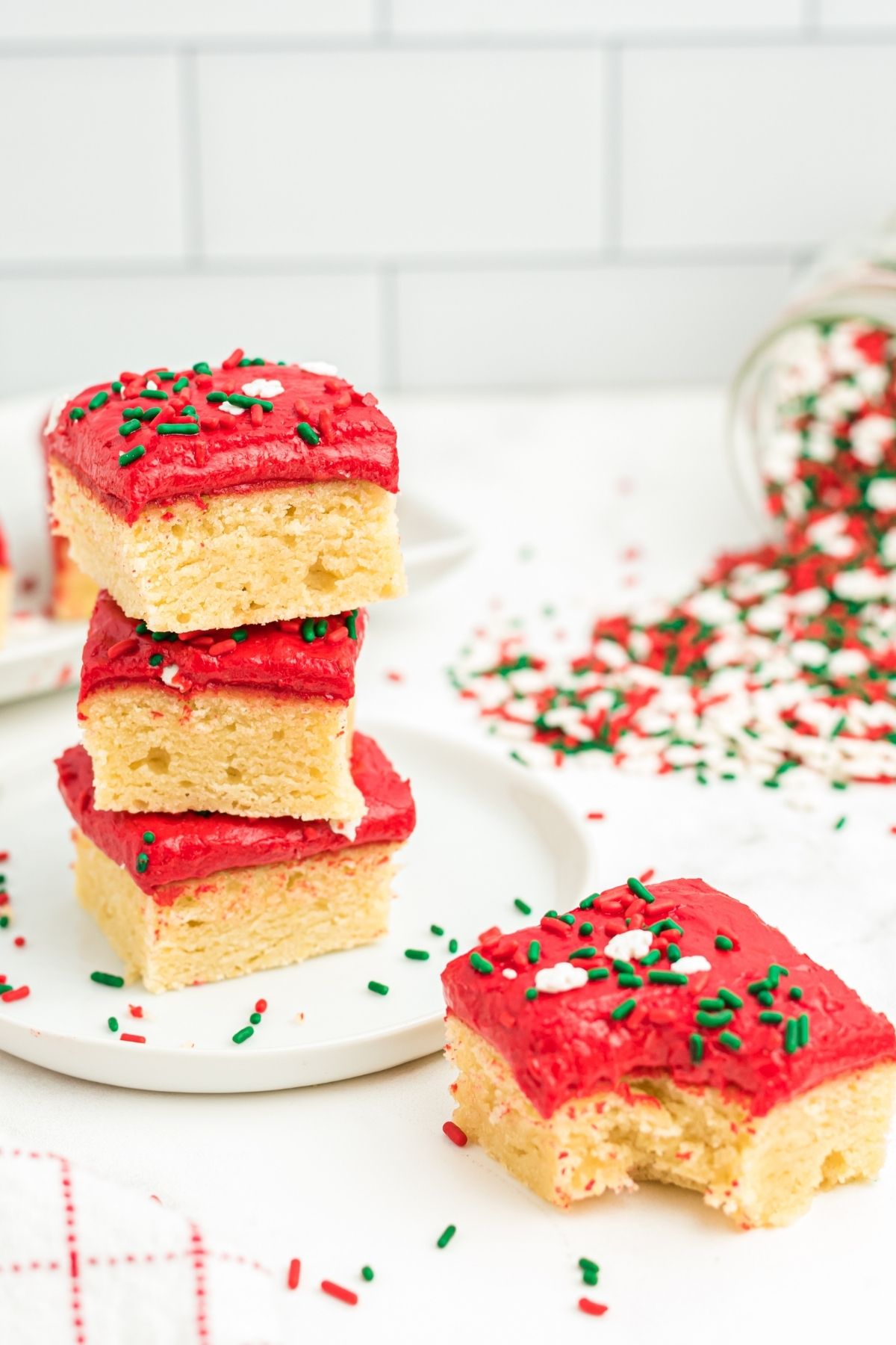 three vanilla sugar cookie bars with red icing and Christmas sprinkles stacked on top of each other, with one with a bite out on the counter