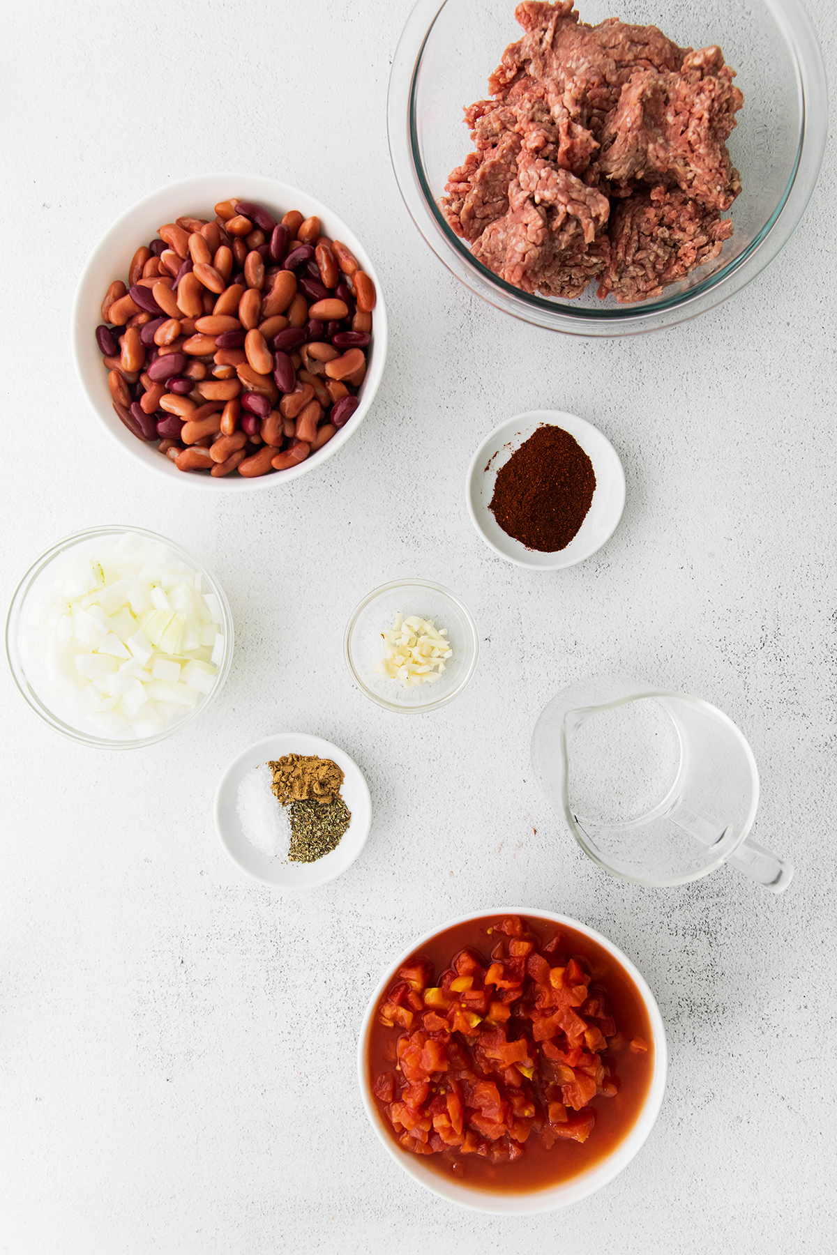 Ingredients on a white counter: beef, yellow onion, garlic, tomatoes, chili powder, kidney beans, cumin, oregano, salt, onion
