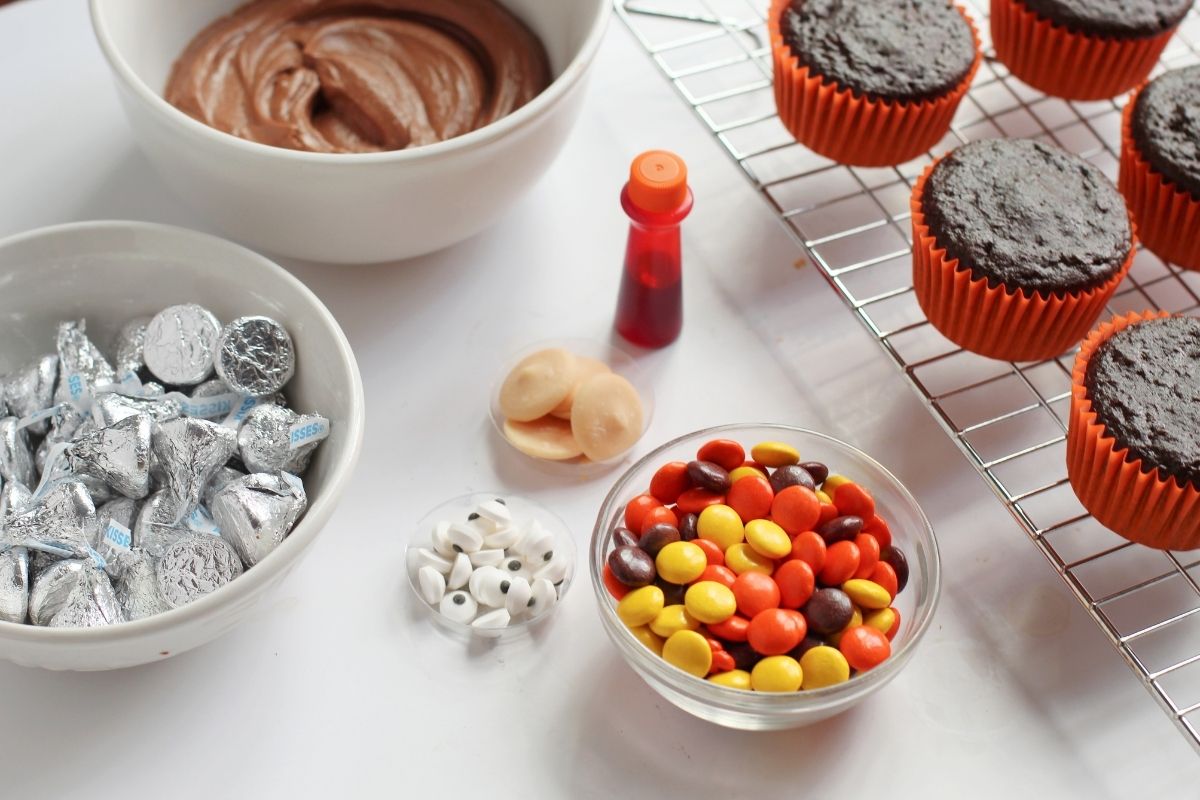 ingredients on white counter: brown frosting, Hershey's kisses, Reese's pieces, chocolate cupcakes in orange wrappers, candy eyes