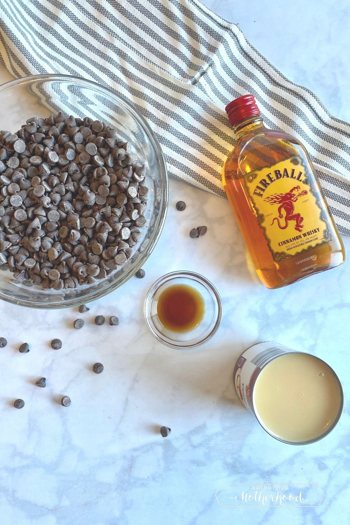 bottle of Fireball whisky, can of sweetened condensed milk (opened), chocolate chips, and vanilla extract on white counter