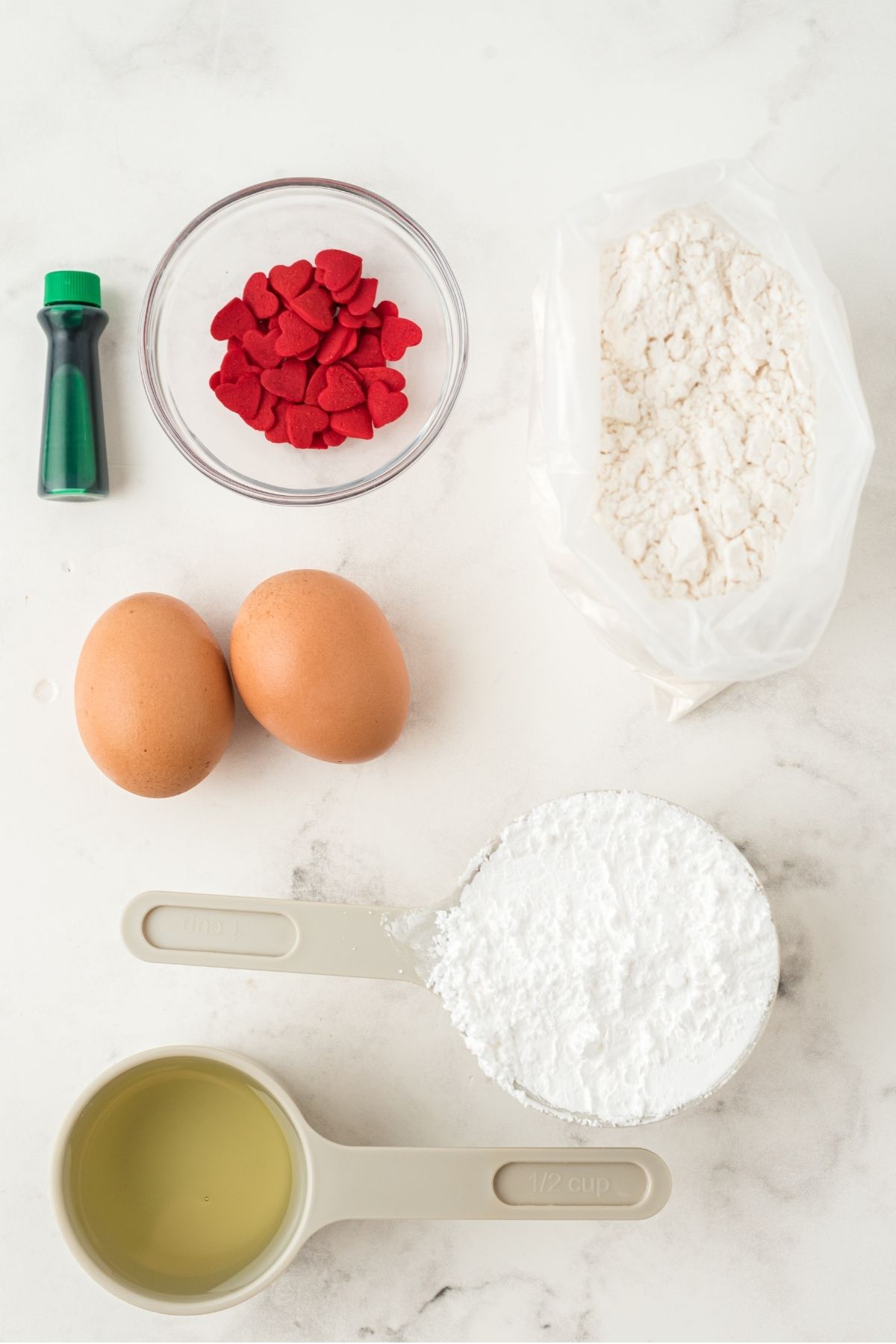 ingredients on white marble counter: green food coloring; red heart sprinkles; white cake mix; 2 brown eggs, confectioner's sugar, vegetable oil