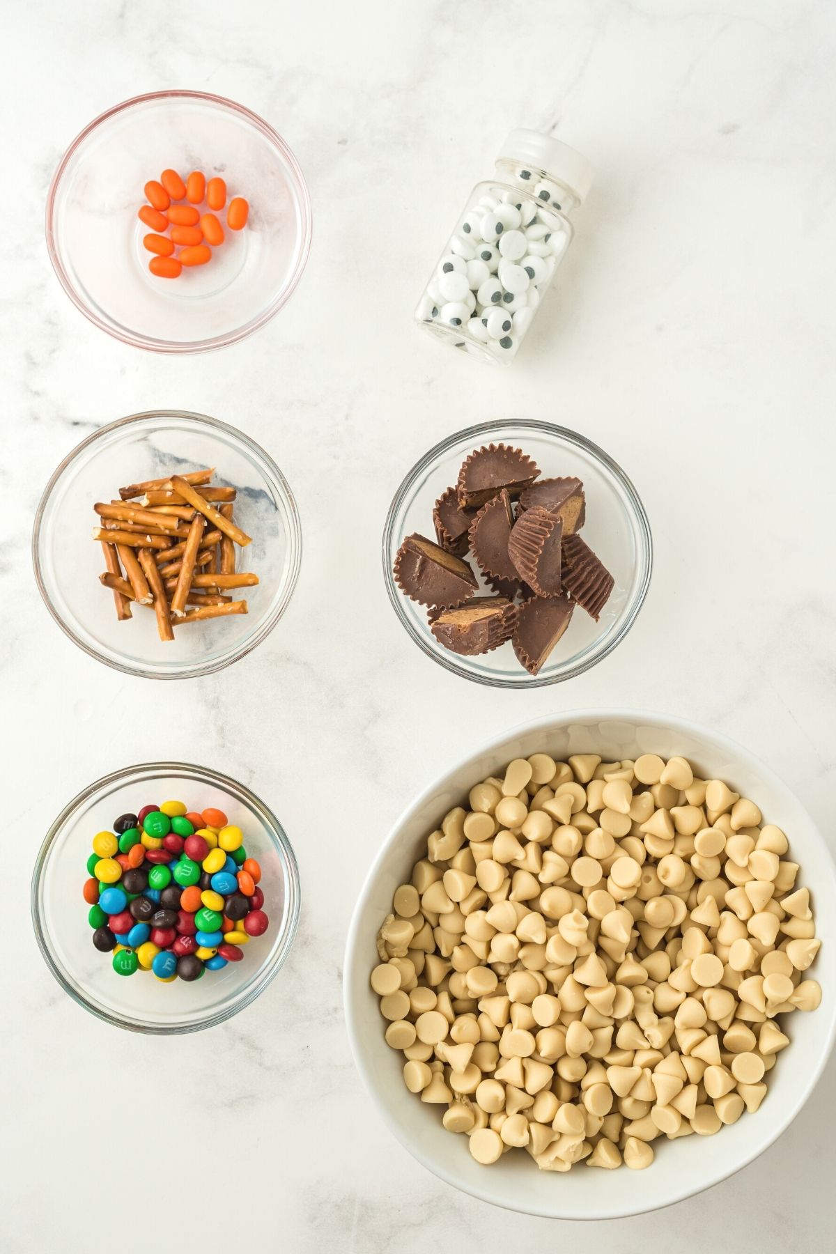 ingredients on white counter: orange tic tacs, candy eyes in a jar; pretzel sticks, Reese's peanut butter cup minis; mini M&Ms; white chocolate chips