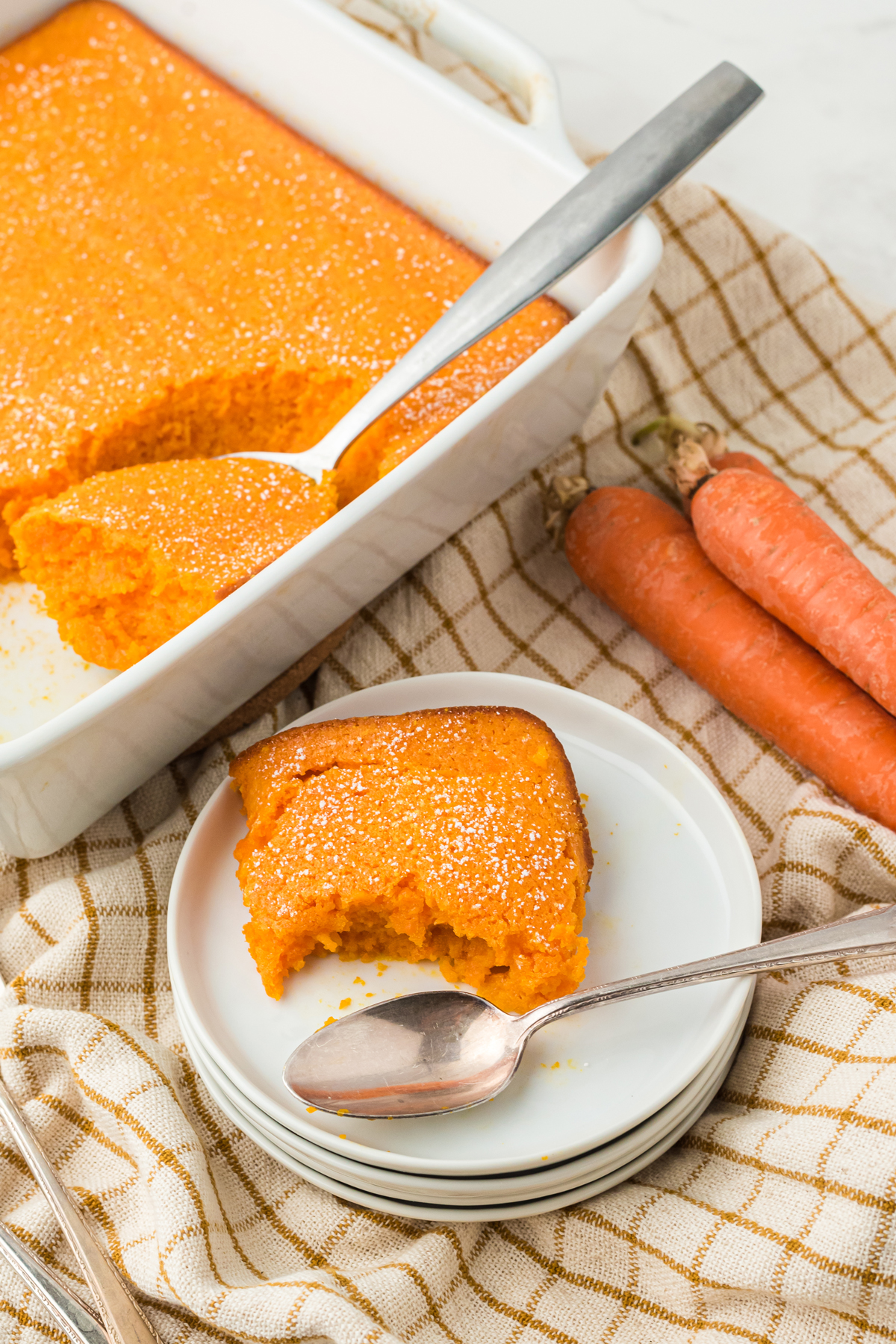 A slice of carrot souffle on a white dessert plate with a spoon. Background is a linen, carrots and a tray of the recipe.