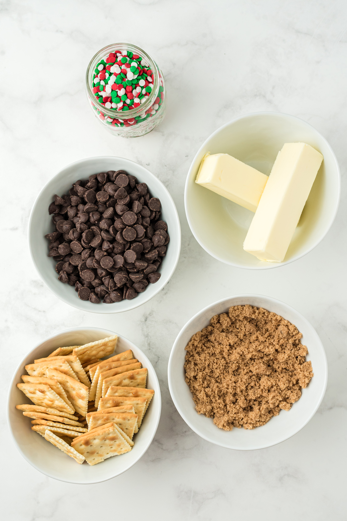 Ingredients of Christmas crack recipe on a white counter. These are crackers, sugar, butter, sprinkles and choco chips