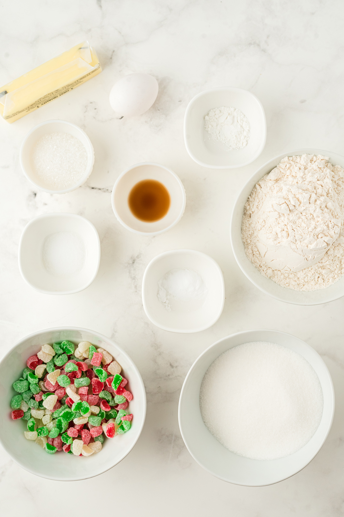Ingredients of Gum Drop Cookies recipe on a white counter. These are cup of all-purpose flour, baking soda, salt, butter, granulated sugar, egg, vanilla extract, greenm red and white-spiced flavored gumdrops, and white sugar.