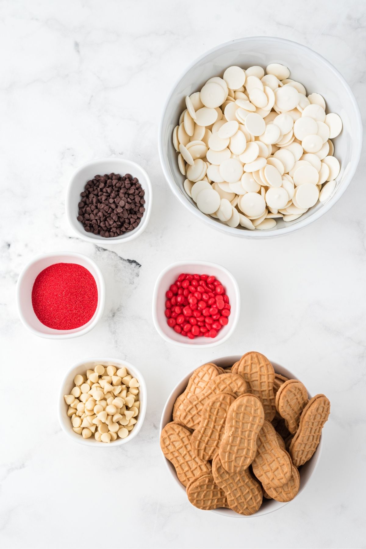 ingredients on white counter: white candy melts, mini chocolate chips, red hot candies, red sanding sugar, Nutter Butter cookies