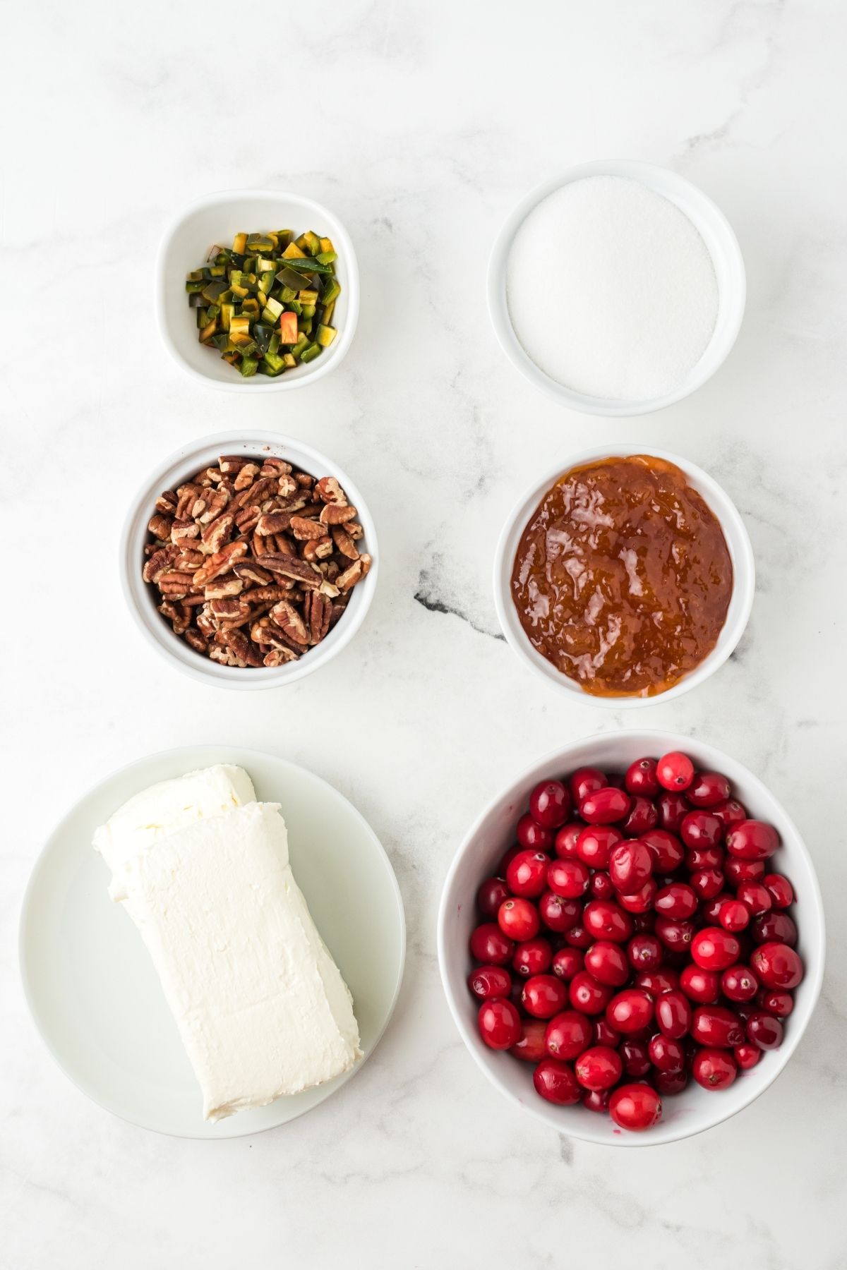 ingredients in bowls on white counter: chopped jalapenos, sugar, apricot jelly, chopped pecans, whole cranberries, block of cream cheese