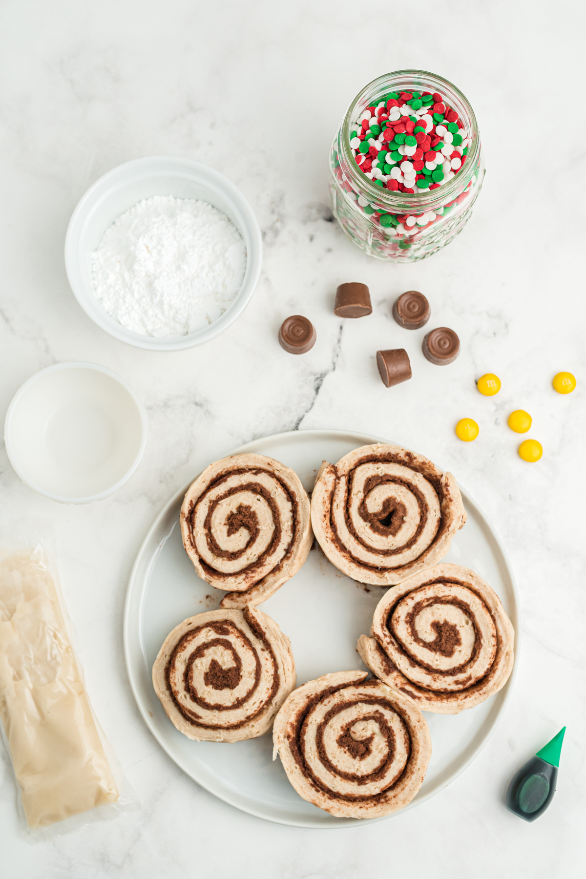 Ingredients of Christmas Tree Cinnamon Rolls on a white counter. These are the following: refrigerated cinnamon rolls, confectioner's sugar, water, green food coloring gel, Christmas sprinkles, yellow M&Ms, miniature peanut butter cups or Rolo candies unwrapped. 