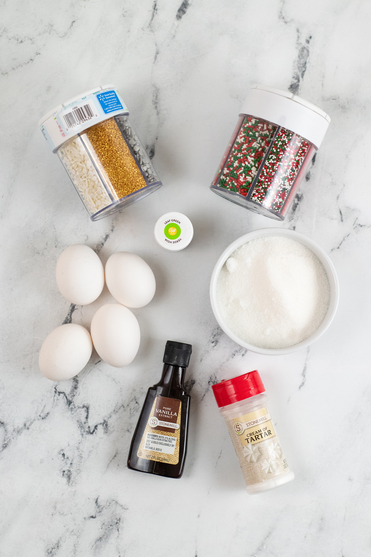 Ingredients for Christmas Tree Meringues on a white counter. This includes eggs, white sugar, vanilla extract, cream of tartar, green gel food coloring, star shaped sprinkles, and sprinkles.
