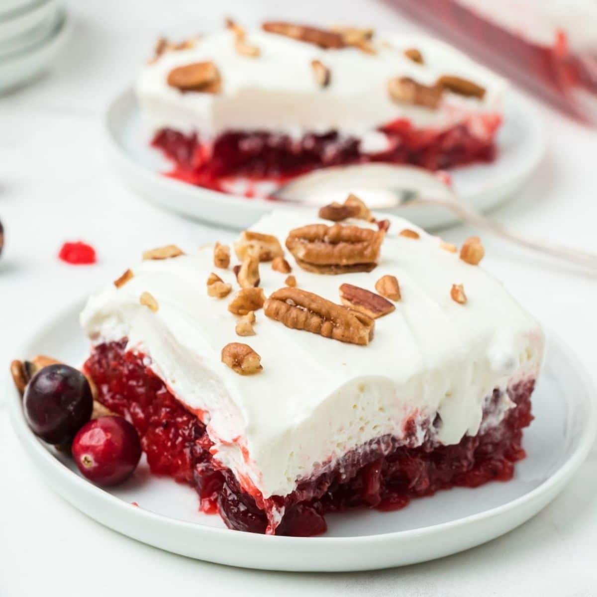 A slice of cranberry jello salad topped with whipped cream and pecans on a white plate, with fresh cranberries on the side.