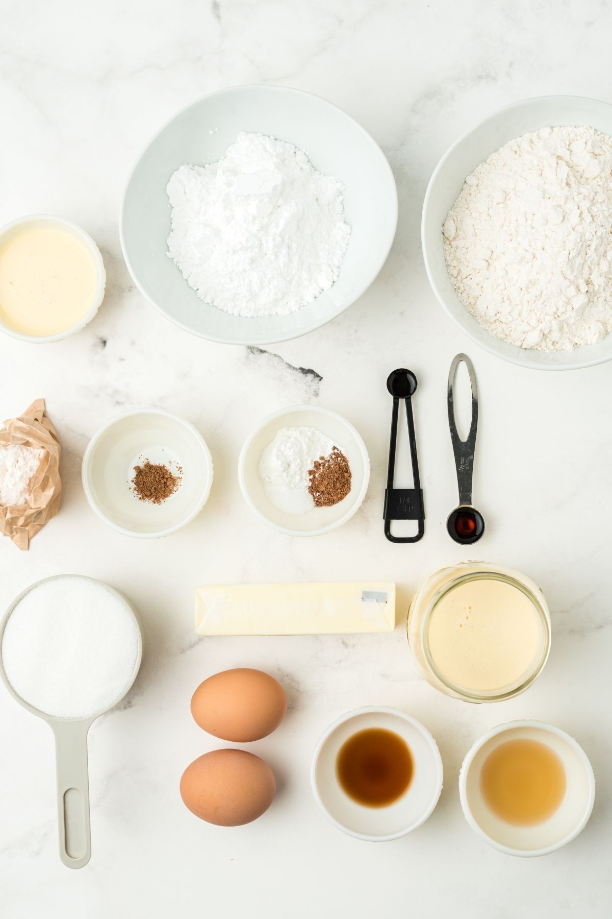 ingredients on white counter: flour, powdered sugar, spices, egg nog, butter, pudding packet, 2 brown eggs, rum extract, vanilla extract, sguar