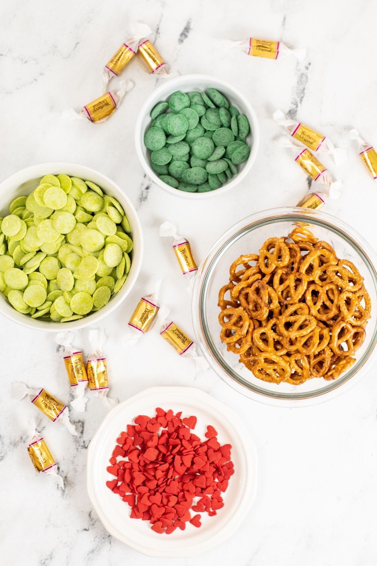 ingredients in bowls on white counter: dark green and light green candy melts, pretzels, red heart sprinkles, caramel candies