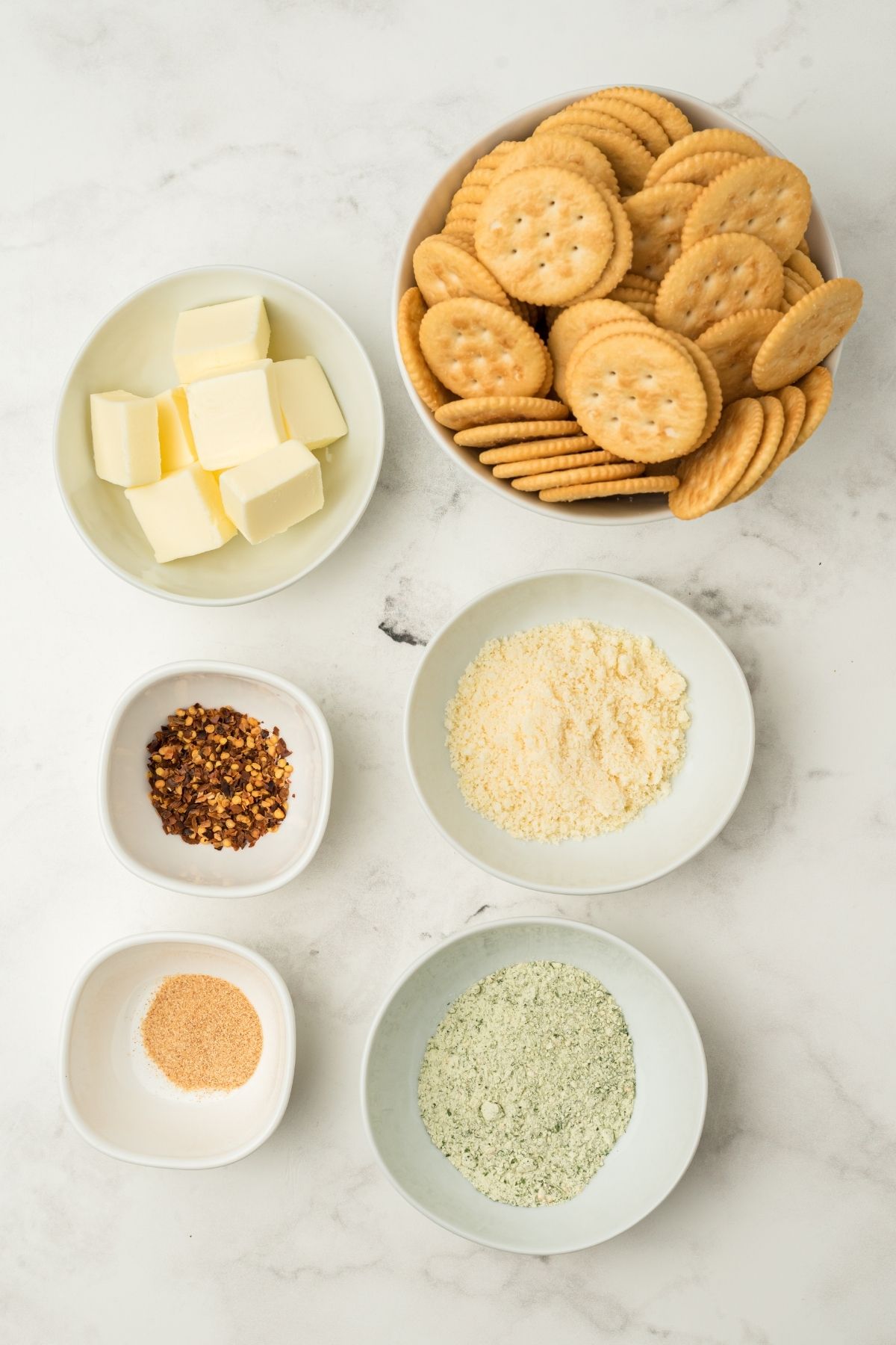 ingredients in bowls on white counter: butter, plain Ritz crackers, parmesan cheese, red pepper flakes, powdered ranch seasoning, garlic powder
