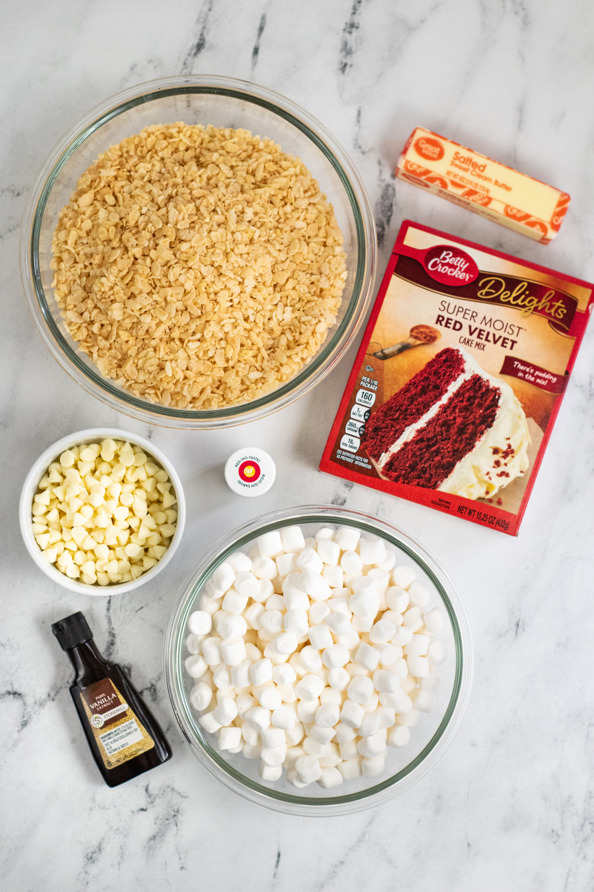 Ingredients in bowls on a marble worktop including: Mini Marshmallows, Butter, Rice Krispies Cereal, Red Velvet Cake Mix, Vanilla Extract, White Chocolate Chips, Taste Free Red Gel Food Coloring