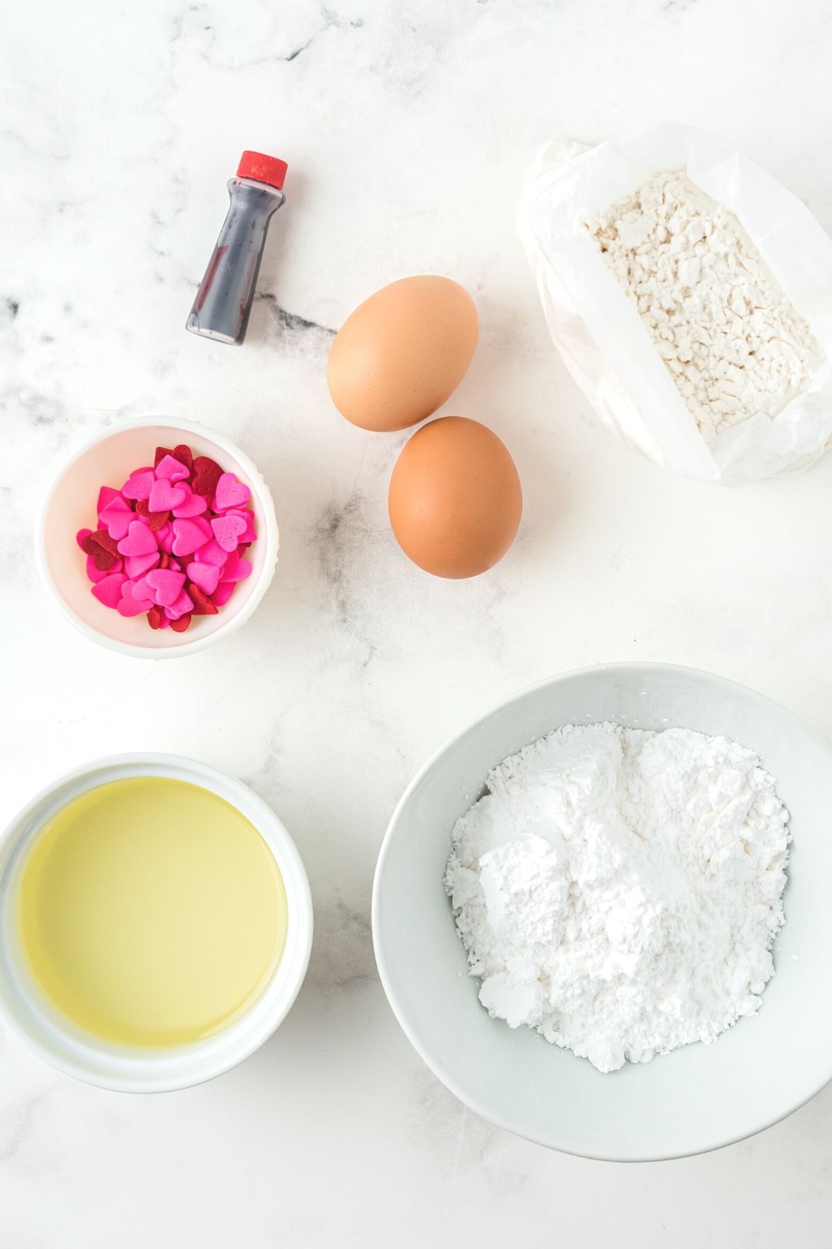 ingredients in white bowls on white marble counter: white cake mix, vegetable oil, eggs, powdered sugar, food coloring, red and pink heart sprinkles