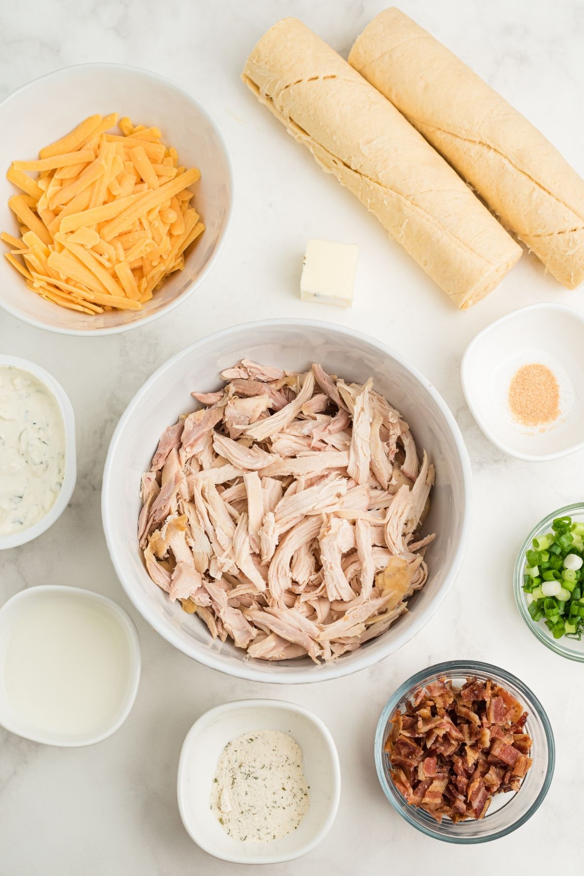 ingredients in white bowls on counter: cheese, shredded chicken, seasonings, cream cheese, Ranch, scallions, chopped bacon and two rolls of crescents