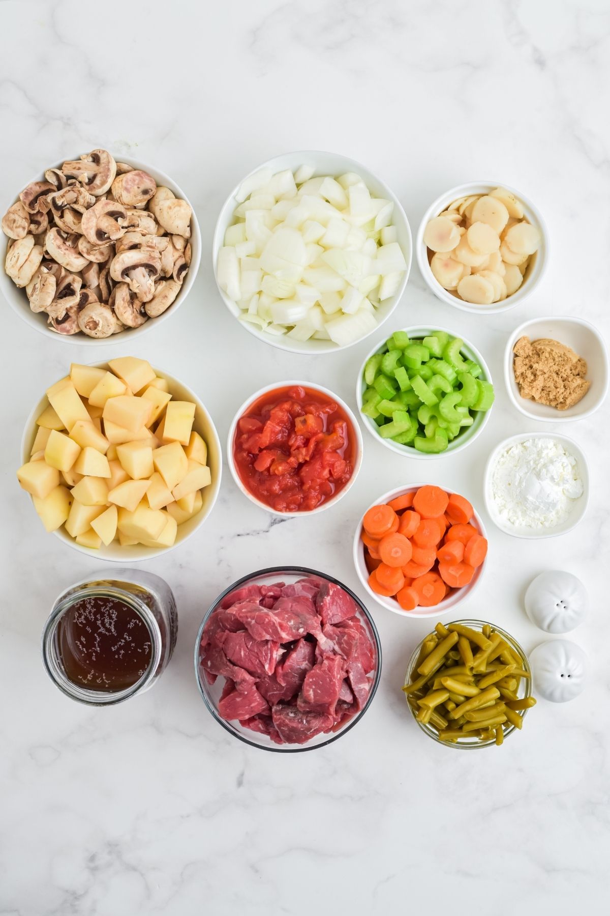 ingredients in white bowls on white counter: mushrooms; onions, water chesnuts, brown sugar, celery, tomato paste, carrots, green beans, potatoes, beef cubes, beef broth